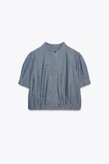 Camisa de denim azul con cuello redondo, manga corta abullonada con pliegues y bajo abullonado con pliegues. Cierre frontal con botones ocultos por solapa.