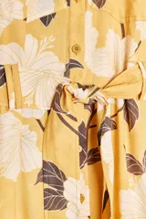 Vestido midi camisero color amarillo con estampado de flores blancas y hojas marrones. Tiene cuello solapa, mangas 3/4 con puños, cinturón de lazo ajustable y cierre frontal con botones.