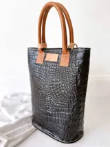 Bolso matero blanco con laterales en cuero negro con textura de cocodrilo, manijas de cuero color suela y accesorio metálico plateado en forma de cruz.