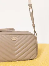 Bandolera color beige con diseño acolchado en forma de V y logo metálico dorado en forma de labios. Tiene correa larga ajustable y herrajes plateados.