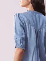 Blusa celeste de lyocell, con cuello a la base, escote en V y botones. Delantera con bolsillo y manga corta con terminación en punto smock.
