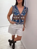 Chaleco corto de denim azul con bordados florales en blanco y marrón, detalle desflecado en escote, sisas y ruedo. Escote en V y cierre frontal con botones. Estilo boho chic.