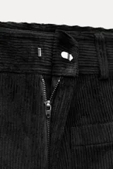 Pantalón ancho de pana color negro, de tiro medio, con trabillas en la cintura, bolsillos delanteros y cierre frontal con cremallera, botón y gancho metálico.