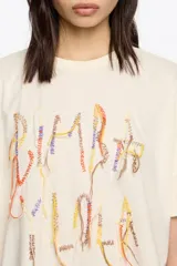 Camiseta oversize de algodón y poliéster color crema con escote redondo y mangas cortas anchas. Presenta un bordado frontal con hilos colgantes de colores variados.