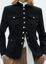 Chaqueta negra con cuello mao y cierre frontal con botones dorados. Tiene cuatro bolsillos de parche con solapa y botón.