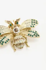 Pin metálico con forma de abeja en acabado dorado, decorado con incrustaciones de cristales tipo strass en colores verde, blanco y marrón.