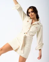 Vestido corto color crema con cuello camisero, cierre frontal con botones, cinturón de la misma tela y mangas largas con puños abotonados. Tiene bolsillos tipo parche en el pecho y costuras a contratono.