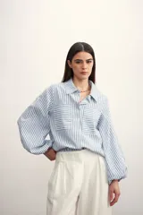 Camisa a rayas verticales color beige y blanco, con cuello clásico, cierre frontal con botón dorado, mangas largas abullonadas con puños elásticos, dos bolsillos frontales y corte holgado.