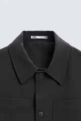 Sobrecamisa negra de corte amplio, con cuello solapa, manga larga con puños abotonados, bolsillos de parche en el pecho, bolsillos laterales y cierre frontal con botones.