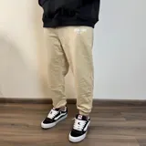 Pantalón de jogging color beige con puños elásticos y estampado con la frase "Buena Vibra Est. 2018" en la parte superior de la pierna izquierda.