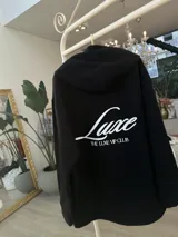 Buzo hoodie negro de corte oversize, con capucha, bolsillo frontal y estampa blanca en la espalda con la leyenda "Luxe" y "The Luxe Vip Club".