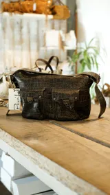 Bolso bandolera color borgoña con textura símil piel de cocodrilo, con correa fina ajustable y dos bolsillos frontales con cierre de broche metálico.