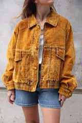 Campera estilo bomber o trucker de color mostaza/naranja quemado con efecto acid wash o desgastado. Presenta cuello camisero, cierre frontal y puños y cintura ajustados con elástico. Posee bolsillos frontales con solapa y botón.