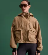 Campera corta color marrón claro, confeccionada en sarga de algodón y poliamida, con cuello alto, capucha y bolsillos delanteros con solapa. Tiene cierre frontal con botones a presión y mangas largas con puños elásticos.