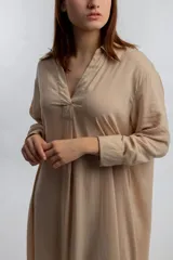 Vestido camisero midi color beige, de corte holgado, con cuello camisero, mangas largas con puños abotonados y aberturas laterales en el ruedo.