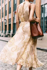 Vestido midi de cuello halter con estampado paisley en tonos beige, amarillo y marrón. Tiene lazada en la espalda, cintura elástica y falda plisada. Cierre trasero con cremallera oculta, abertura y botón.