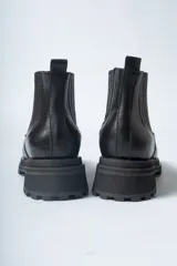 Bota corta tipo Chelsea de cuero negro, con puntera cuadrada y suela track gruesa y dentada. Presenta paneles elásticos acanalados a los lados para facilitar el calce.