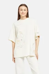Remera color crema de corte holgado con cuello redondo y mangas cortas. Presenta un estampado floral en tonos beige y marrón a lo largo del frente y la manga derecha.