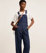 Jardinero largo de jeans azul oscuro, con breteles anchos ajustables con botones, bolsillo tipo parche en el pecho y cierre lateral con botones.