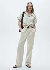 Blusa corta de manga corta con diseño a cuadros en tonos beige y blanco. Presenta cuello camisero, cierre frontal con botones y lazo en la parte inferior.