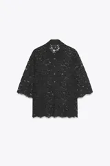 Camisa semitransparente de encaje negro con cuello solapa, manga tres cuartos y bajo festoneado. Cierre frontal con botones metálicos en forma de flor.