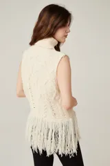 Chaleco de punto sin mangas color beige, con cuello alto tipo polera. Presenta un diseño de tejido con ochos y rombos, y terminación de flecos largos en el ruedo.
