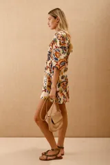 Vestido corto estampado con motivos florales en tonos naranja, azul y verde sobre fondo blanco. Tiene mangas 3/4 con volados en los puños, escote en V y cintura elastizada con lazo.