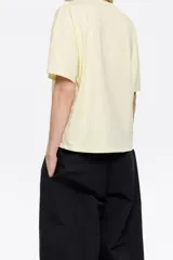 Camiseta color crudo de algodón, con cuello redondo y mangas cortas. Presenta logo bordado en color dorado en el frente.