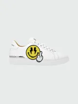 Zapatillas bajas de cuero blanco con aplique de parche bordado de cara sonriente amarilla con relámpagos en los ojos y dedo medio levantado.