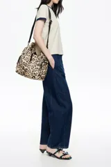 Bolso bandolera de estructura blanda marca Bimba y Lola, confeccionado en nylon con estampado animal print de leopardo. Presenta doble asa de mano integrada, correa de tejido negra regulable y extraíble, bolsillos laterales con cierre y logo metálico de la marca en el frente.