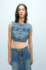 Chaleco crop de denim celeste, sin mangas, con cuello redondo, cierre frontal con botones metálicos, dos bolsillos con solapa en el pecho y corte ajustado.