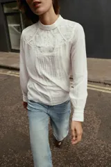 Blusa blanca de cuello redondo y manga larga con puños. Presenta bordados florales y entredoses combinados en el frente. Cierre en la espalda con abertura y botón.