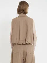 Chaleco marrón de gabardina con corte recto y estilo utilitario. Tiene cierre frontal con tirador de flecos largos en cuero y bolsillos grandes con tapa en el frente.