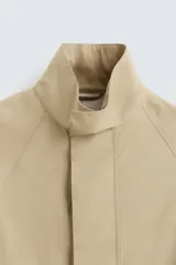 Parka ligera color beige, de corte relaxed fit, confeccionada en tejido técnico repelente al agua. Tiene cuello alto, mangas largas con puños ajustables con botón a presión, bolsillos de solapa en la cadera y cierre frontal con cremallera oculta por solapa con botones a presión.