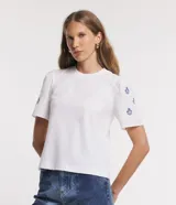 Remera blanca de algodón con cuello redondo, mangas cortas abullonadas y pequeños bordados florales azules en las mangas.