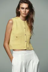Chaleco amarillo de punto con cuello redondo y sisas amplias. Tiene cierre frontal con botones dorados y dos bolsillos de parche con botón.