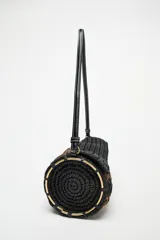 Bolso de hombro con cuerpo trenzado en color negro y marrón. Tiene detalle metálicos circulares en los laterales, asa de hombro, bolsillo interior con cremallera y cierre mediante solapa y cremallera.