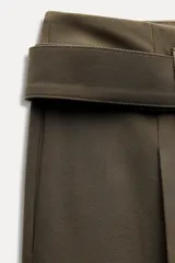 Pantalón de tiro medio color verde oliva, con cintura con trabillas, bolsillos laterales ocultos, cinturón ajustable con hebilla metálica, pinzas delanteras y cierre frontal con cremallera, botón y gancho metálico.