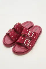 Sandalia tipo birkenstock de color rojo oscuro o bordó, con dos tiras anchas ajustables con hebillas metálicas plateadas. Las tiras están adornadas con apliques metálicos redondos (ojales). La suela es gruesa y del mismo color que la capellada.