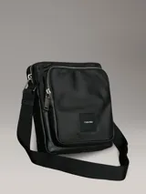 Bolso tipo reportero o bandolera Calvin Klein de color negro, con cuerpo principal de tejido texturizado y detalles en material liso en los bordes. Presenta un bolsillo exterior con cierre de cremallera en la parte superior y un parche cuadrado con el logo de Calvin Klein en la parte frontal inferior.