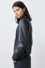 Chaqueta corta de cuero sintético color negro, con cuello redondo y cierre frontal. Presenta detalle de pasamanería en hombros y cuello.