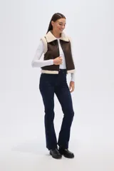 Chaleco sin mangas de eco-cuero marrón con forro y cuello de piel sintética color crema. Presenta cierre frontal y detalles de hebillas ajustables en los laterales.