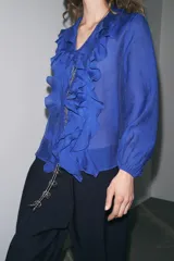 Camisa azul con escote en pico, cierre frontal con botones, mangas largas abullonadas con elástico en los puños y detalle de volantes en el frente.