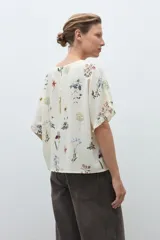 Blusa de manga corta, color crudo, con estampado floral en tonos azules, amarillos y rojos. Presenta cuello redondo y volados en los puños.