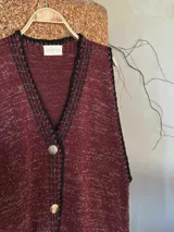 Chaleco de punto color borgoña con detalles de lurex dorado, escote en V, terminaciones en crochet negro y cierre frontal con botones plateados.