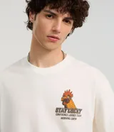 Remera color crudo de corte boxy, con cuello redondo y mangas cortas. Estampado trasero con un gallo con gafas y zapatillas, la palabra 'Rooster' y la frase 'Stay Cocky'.