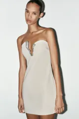 Vestido corto color crema, de escote asimétrico y sin mangas. Presenta una pieza metálica con apliques de strass en el escote.