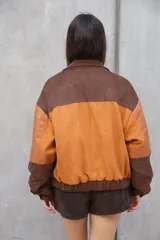 Chaqueta estilo bomber de gamuza con bloques de color en tonos tierra: naranja quemado, marrón oscuro y marrón claro. Presenta detalles en cuero liso en los hombros y bolsillos frontales con tapa y broche dorado. El puño y la cintura son ajustados con elástico.