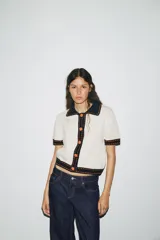 Chaqueta color blanco de punto con cuello solapa y manga corta. Tiene cierre frontal con botones color naranja y ribete en azul marino y naranja.