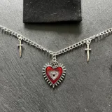 Gargantilla plateada con cadena de eslabones, dije central de corazón rojo con ojo y rosa, y dos dijes de dagas a los lados.